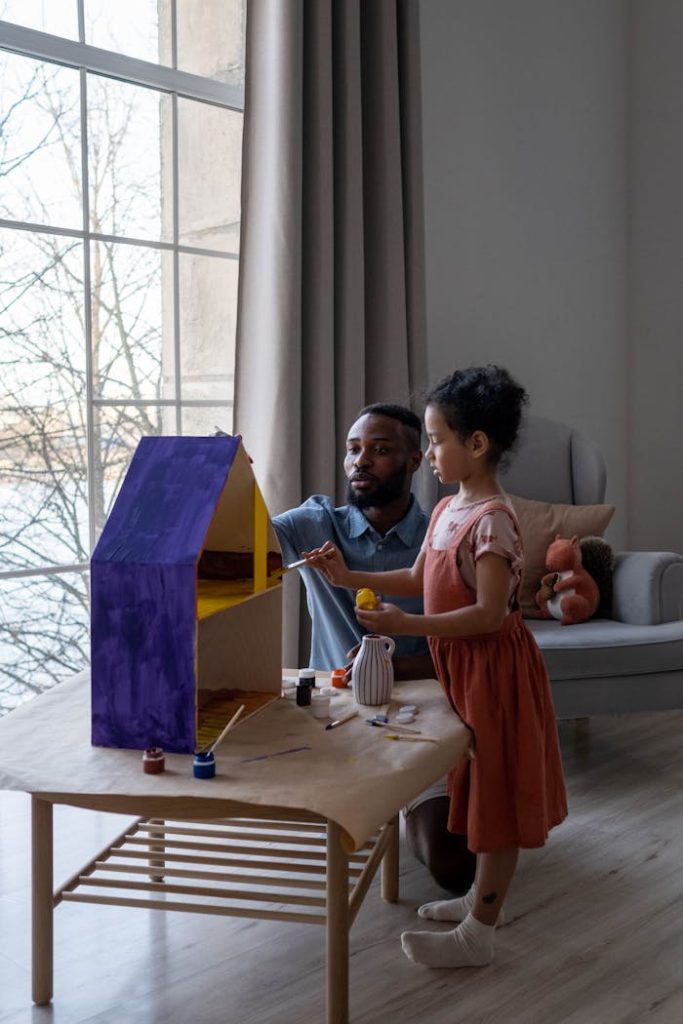 A father and daughter enjoy painting a dollhouse indoors, spending quality family time together.